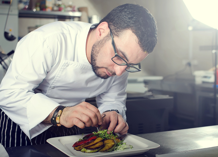 Chef wearing glasses plating a gourmet-style entree featuring fresh grilled vegetables in a professional kitchen setting.