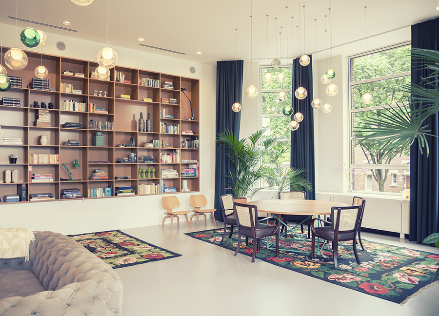 Home library featuring large open windows with window seat, table, plush couches and a large bookcase for displaying books and trinkets.