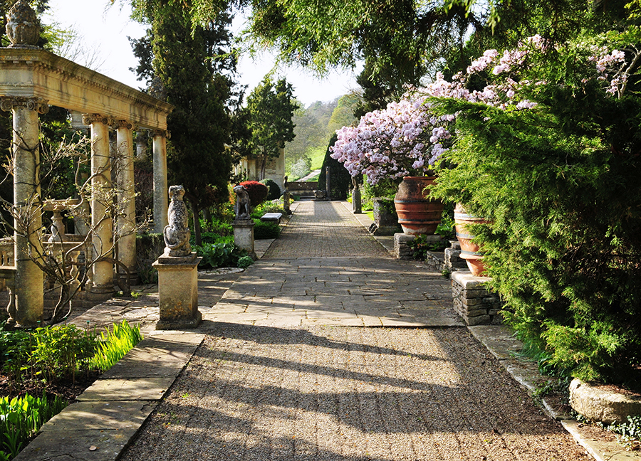 Elegant outdoor garden with stonework, a variety of florals and greenery, statues, and a marble pergola